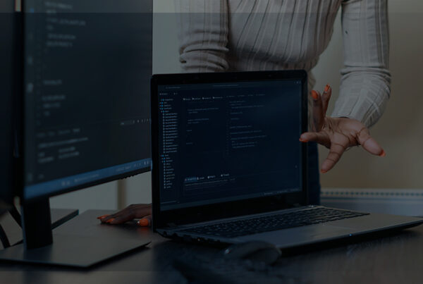 Uma mulher com uma blusa rosa apontando para dois monitores de computador com programas de digitação de códigos de programação | Como calcula a vida útil de um equpamento de TI.
