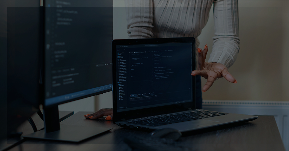 Uma mulher com uma blusa rosa apontando para dois monitores de computador com programas de digitação de códigos de programação | Como calcula a vida útil de um equpamento de TI.