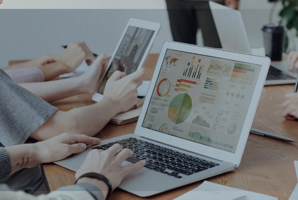 Imagem de uma mesa com vários computadores com telas em gráficos e pessoas em volta trabalhando, representando as transações financeiras empresariais, foco das Fintechs.