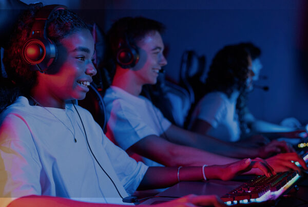 Grupo de jovens jogando em computadores em um ambiente escuro, com luzes coloridas, representando o impacto da tecnologia e telecomunicações no entretenimento.