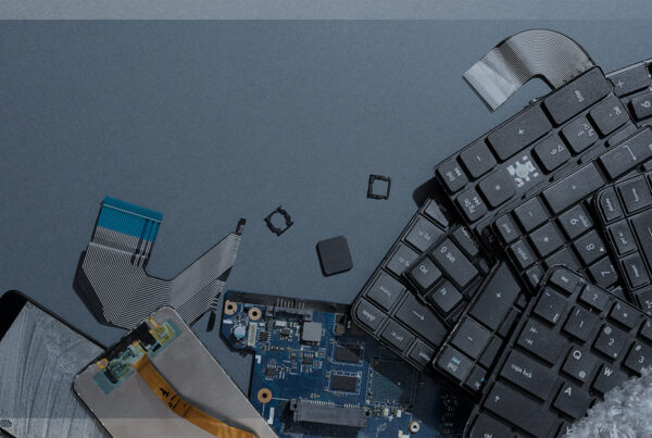 Aparelhos eletrônicos desmontados e quebrados, incluindo teclado, placa-mãe, tela de celular e cabos flat, simbolizando o descarte ou reciclagem de componentes eletrônicos.