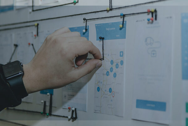 Mão ajustando fios conectados a alfinetes em um quadro com mapas e fluxos de trabalho, simbolizando análise e planejamento estratégico.