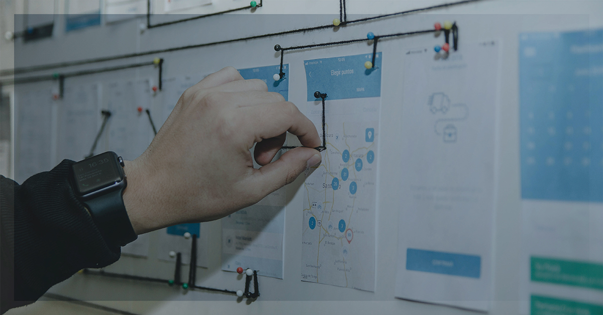 Mão ajustando fios conectados a alfinetes em um quadro com mapas e fluxos de trabalho, simbolizando análise e planejamento estratégico.