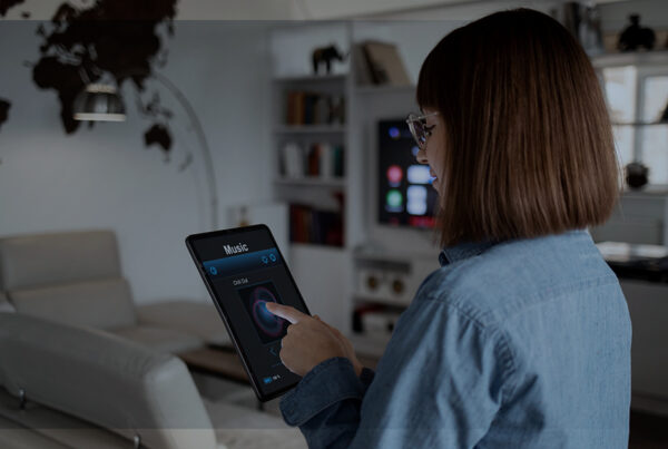 Mulher de costas utilizando um tablet em uma sala moderna, controlando a reprodução de música. Ao fundo, uma TV conectada e elementos de automação residencial, simbolizando uma casa inteligente e integrada.