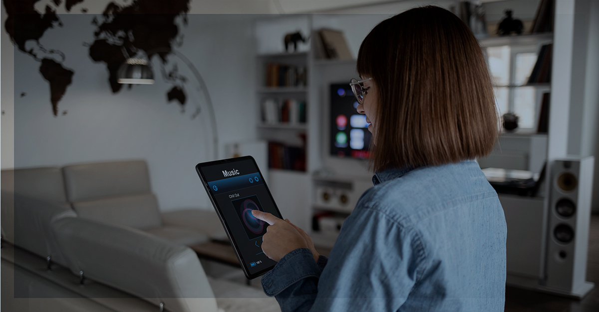 Mulher de costas utilizando um tablet em uma sala moderna, controlando a reprodução de música. Ao fundo, uma TV conectada e elementos de automação residencial, simbolizando uma casa inteligente e integrada.