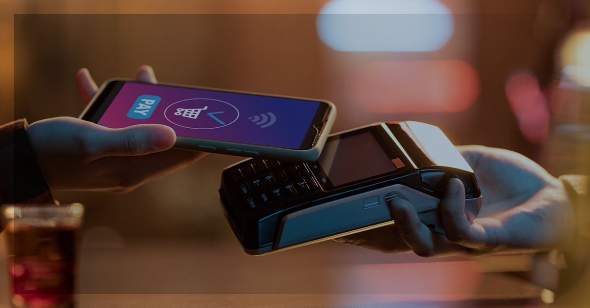 Pessoa aproximando um smartphone de uma máquina de pagamento com tecnologia de aproximação, representando o uso de carteiras digitais para transações sem contato.