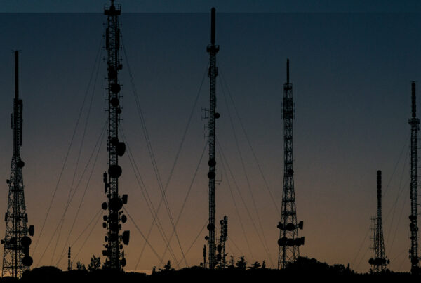 Várias torres de telecomunicação e antenas em silhueta contra o pôr do sol, representando a infraestrutura das redes móveis e antenas inteligentes.