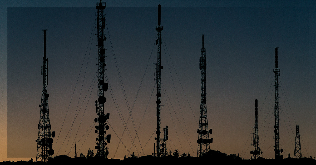 Várias torres de telecomunicação e antenas em silhueta contra o pôr do sol, representando a infraestrutura das redes móveis e antenas inteligentes.
