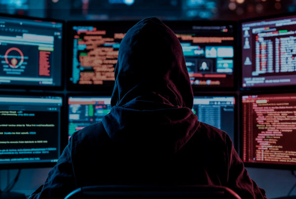 Pessoa com capuz sentada em frente a vários monitores cheios de códigos e gráficos, representando ataques cibernéticos e ataques DDoS a servidores.