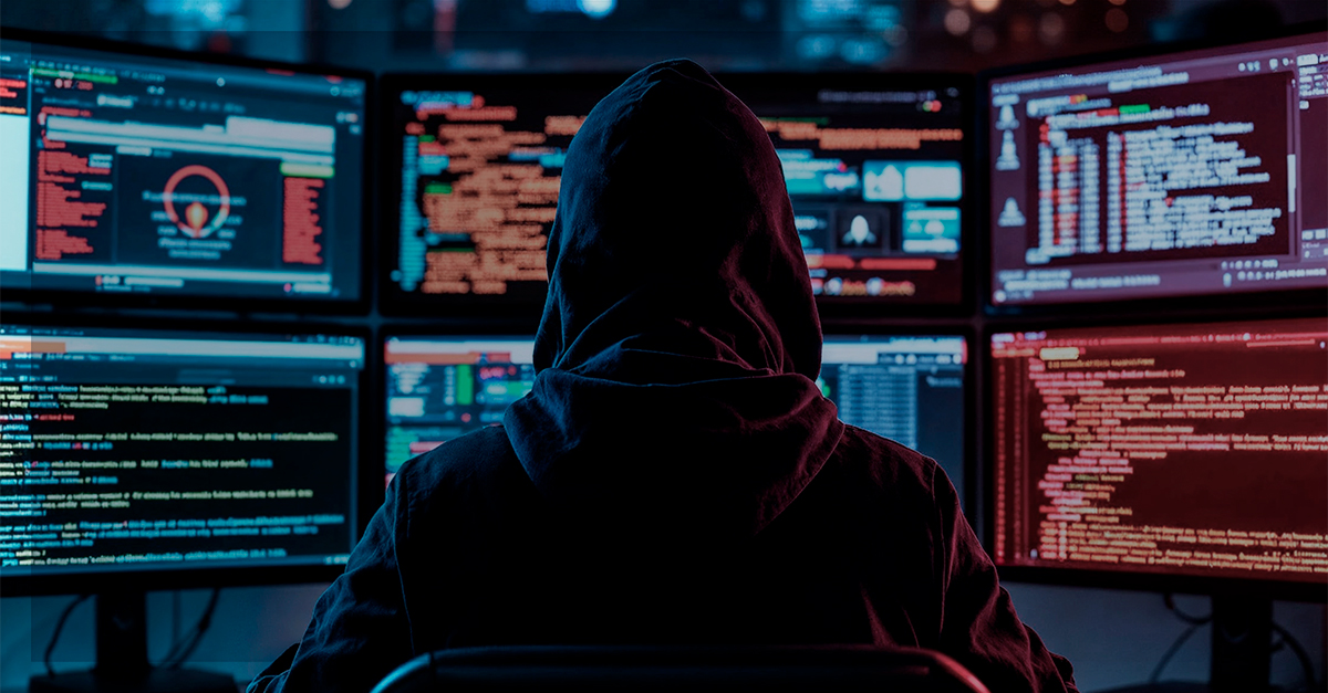 Pessoa com capuz sentada em frente a vários monitores cheios de códigos e gráficos, representando ataques cibernéticos e ataques DDoS a servidores.